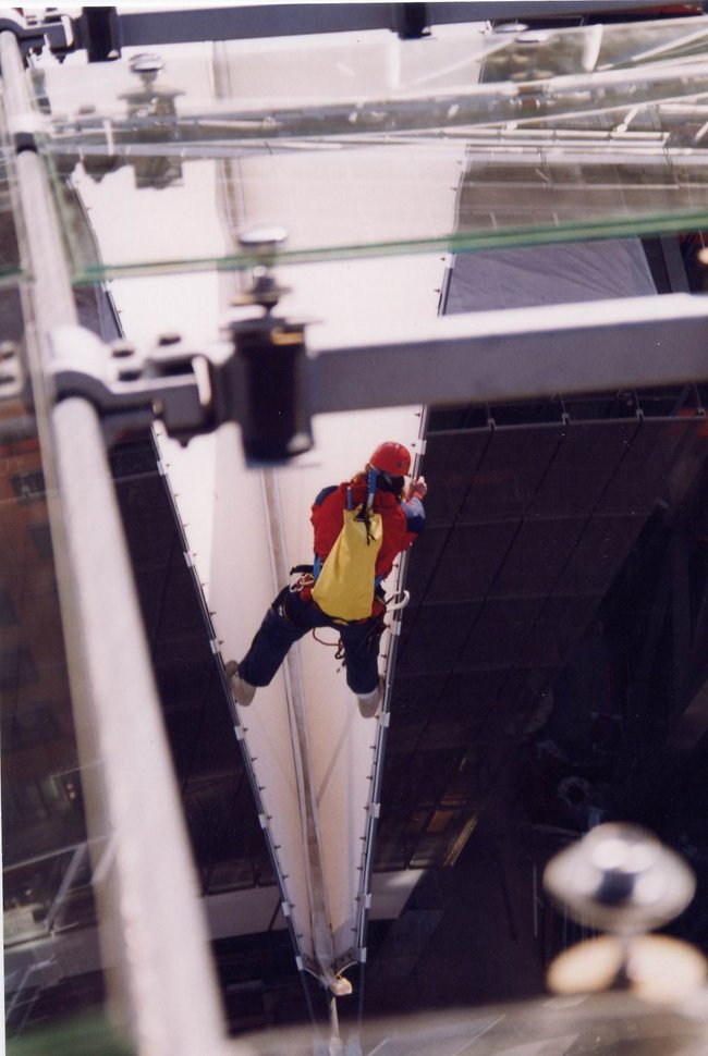 Industriekletterer mit rotem Helm und Sicherheitsgurt beim Klettern an einer Wand, ausgestattet mit gelbem Beutel mit blauen Griffen und Kletterausrüstung. Die Person hält dabei einen Stab.