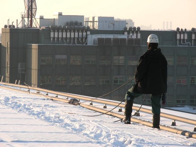 Ein Industriekletterer bei Winterdienstarbeiten auf einem verschneiten Dach, ausgestattet mit schwarzer Schutzkleidung, Helm und Seil, hält ein Werkzeug oder eine Schlauchleitung.