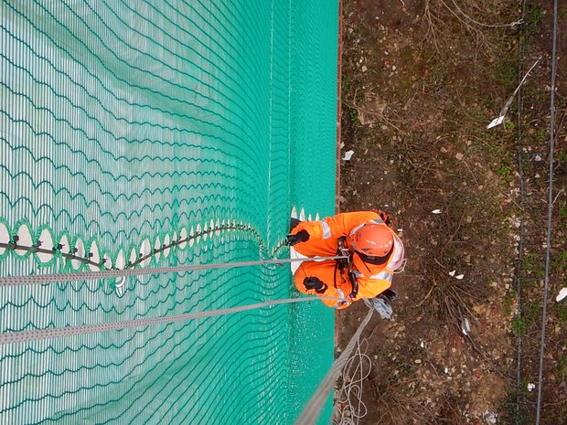 Ein Industriekletterer von ZITRAS in orangen Sicherheits-Overalls und Helm arbeitet an einem grünen Fassadenschutznetz, vermutlich in Duisburg.