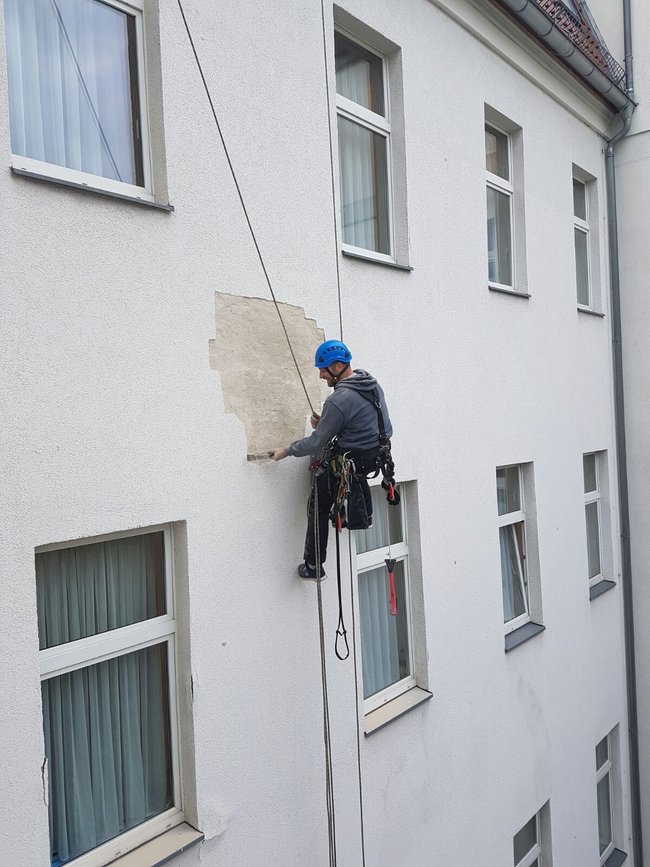 Ein Industriekletterer von ZITRAS in Ausrüstung, mit Helm und Gurt, führt an einem Seil hängend Fassadensanierungsarbeiten an einem Gebäude aus. Im Hintergrund ist ein Fenster mit weißen Rahmen und Vorhängen sichtbar.
