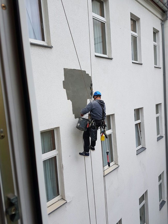 Ein Industriekletterer von ZITRAS führt Verputzarbeiten an einer Gebäudefassade durch. Er ist mit einem Sicherheitsgurt ausgestattet, hält einen Eimer und klettert geschickt an der Wand entlang. Im Fokus stehen eine nah gelegene Fensterfront und die präzise Handarbeit des Kletterers.
