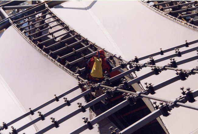 Ein Industriekletterer von ZITRAS arbeitet an einer Metallstruktur, möglicherweise während der Montagearbeiten am Sony Center. Der Kletterer ist mit Sicherheitsausrüstung ausgestattet, trägt einen gelben Beutel und steht auf einem Dach. Außerdem sind ein Helm und Teile der Metallstruktur im Detail erkennbar. Ein schwarzes Objekt im Bild ist unscharf.