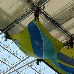 Ein Industriekletterer von ZITRAS bei der Arbeit in großer Höhe unter einem Glasdach, der möglicherweise eine große, grün-blaue Plane oder Schutzabdeckung handhabt