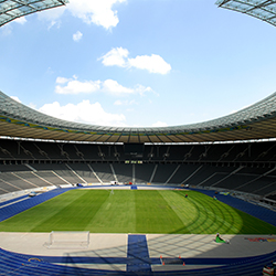 Ein großes Stadion mit grünem Spielfeld und blauen Sitzen mit dem Olympiastadion im Hintergrund