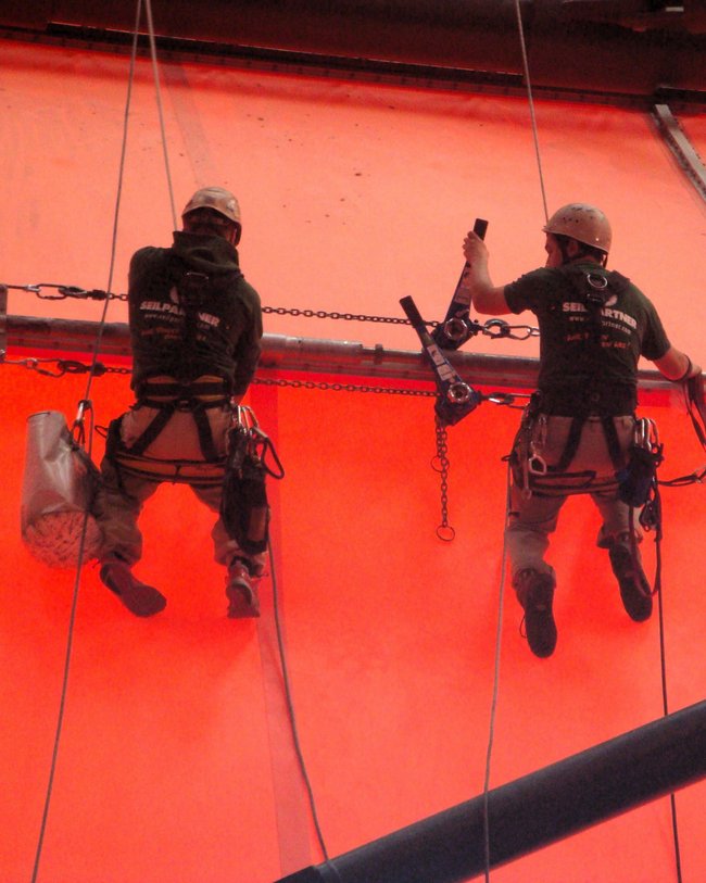 Zwei Arbeiter in Kletterausrüstung arbeiten an einer Kette vor einem orangefarbenen Hintergrund.