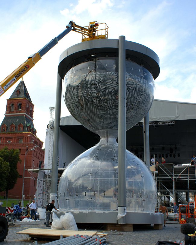 Große Sanduhrinstallation mit transparenten Glaskapseln, umgeben von Gerüsten und einem Kran.