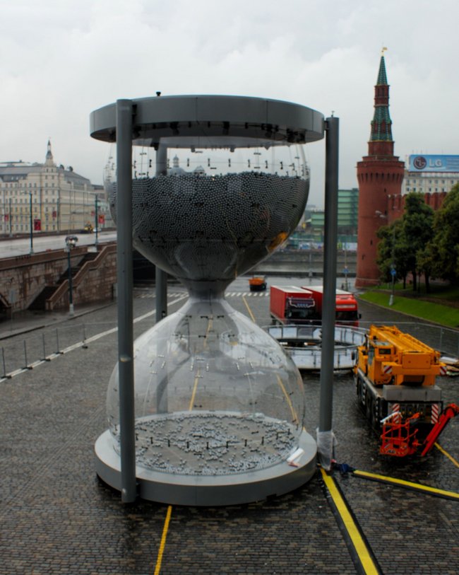 Große Sanduhr aus Metall und Glas in einem urbanen Umfeld mit einem historischen Turm im Hintergrund.