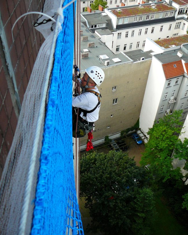 Ein Arbeiter mit Schutzhelm und Sicherheitsausrüstung befestigt ein blaues Netz an einem hohen Gebäude.