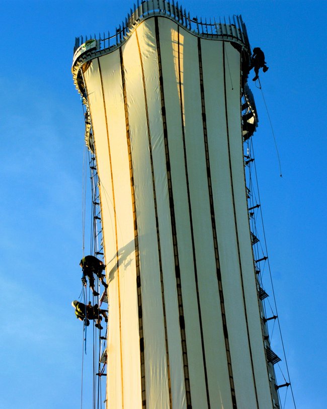 Hoher Aussichtsturm mit Arbeitern, die an der Außenfassade arbeiten, umgeben von blauem Himmel.