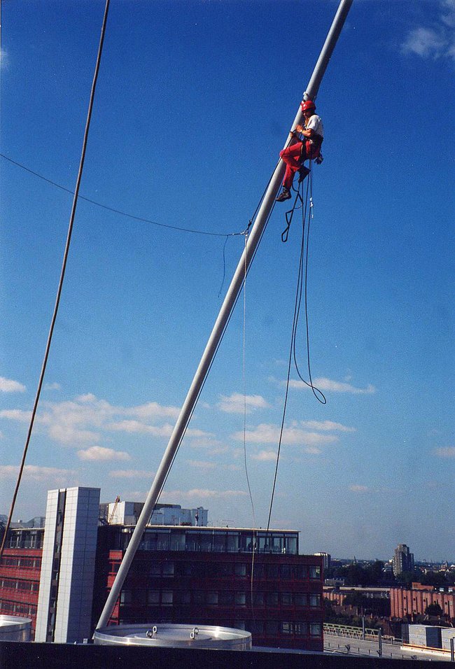 Arbeiter in orangefarbener Sicherheitskleidung klettert an einem schrägen Mast hoch, mit Seilen gesichert, vor blauem Himmel und städtischer Kulisse.