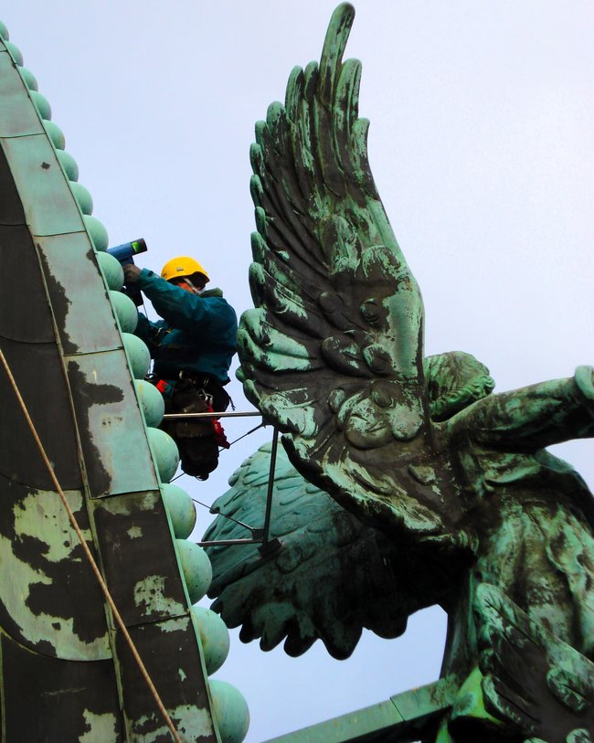 Ein Arbeiter in Schutzkleidung und gelbem Helm, gesichert mit einem Seil, ist auf einem Gerüst hoch oben an der Fassade des Berliner Doms zu sehen. Neben ihm ist eine große, detaillierte Statue mit Flügeln.