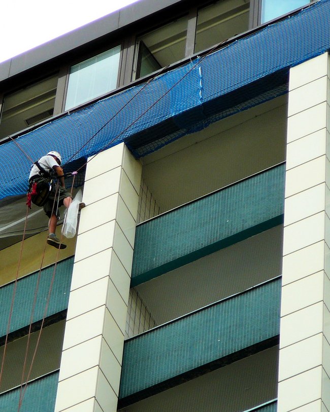 Eine Person in Arbeitskleidung klettert an einem hohen Gebäude und führt Arbeiten an der Fassade durch.