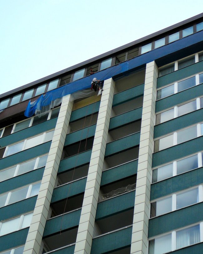 Mehrstöckiges Hochhaus mit zwei Arbeitern, die oben an der Fassade auf einem Gerüst arbeiten.
