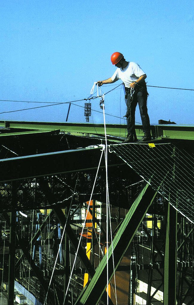 Eine Person mit rotem Schutzhelm und weißem T-Shirt steht auf einer grünen Metallstruktur und bedient ein Seil.