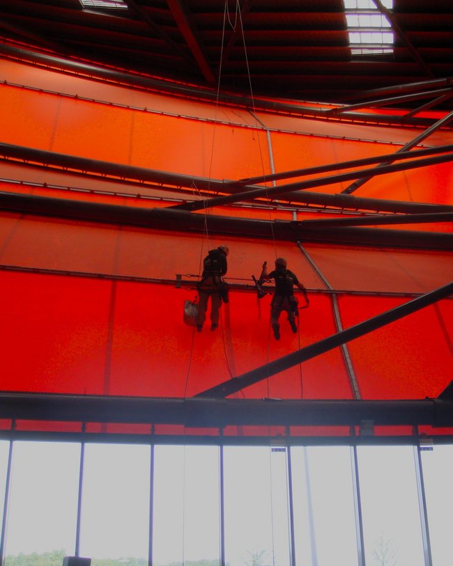 Zwei Personen bei der Arbeit an den Seilen im Inneren einer großen Halle mit leuchtend orangefarbener Decke.