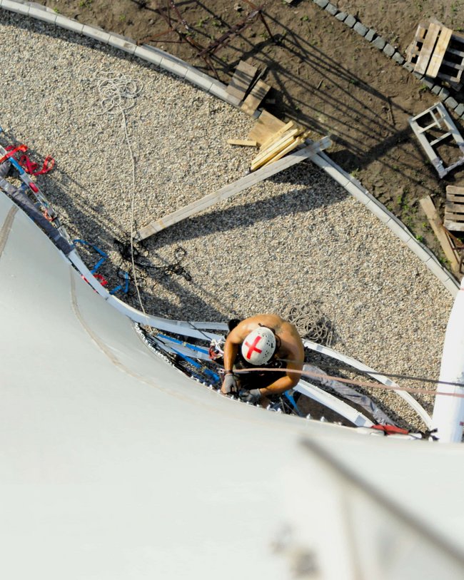 Eine Person mit Helm und Sicherheitsausrüstung klettert an einem Turm nach oben. Blick von oben auf die Person.