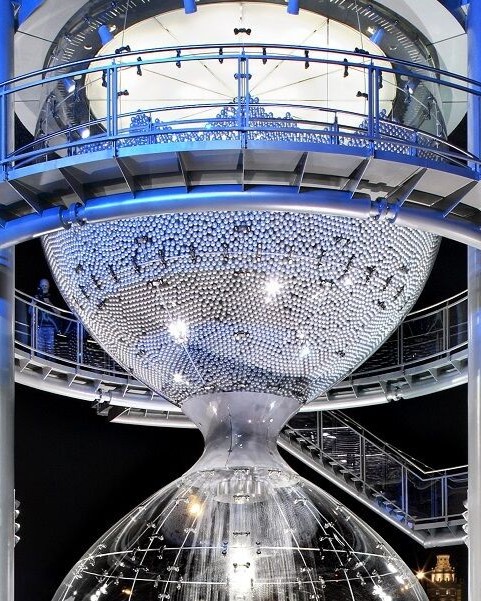 Große, futuristische Sanduhr mit transparenter Mittelachse und reflektierenden Metallkugeln oberhalb und unterhalb der Glasmitte.